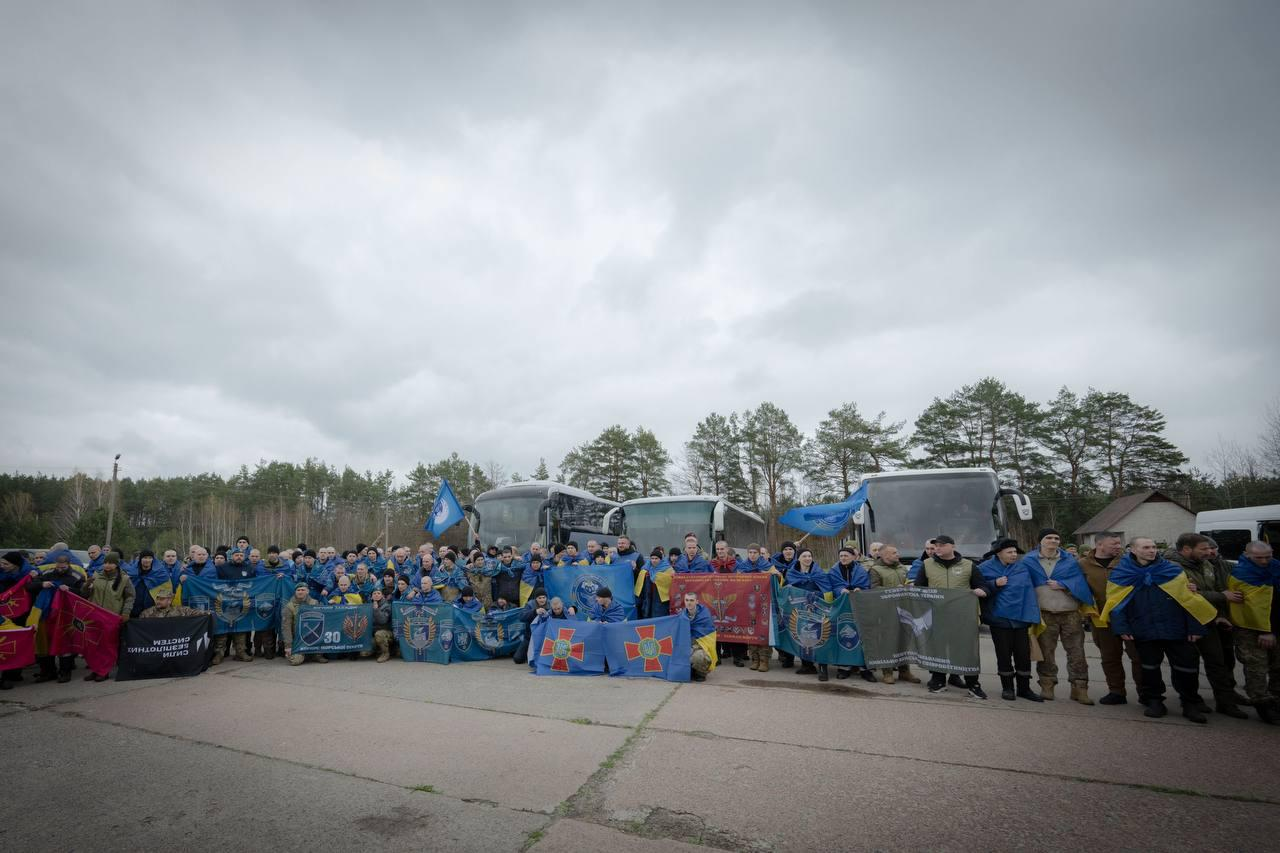 Група звільнених захисників України стоїть на дорозі біля автобусів під похмурим небом, тримаючи державні та військові прапори.