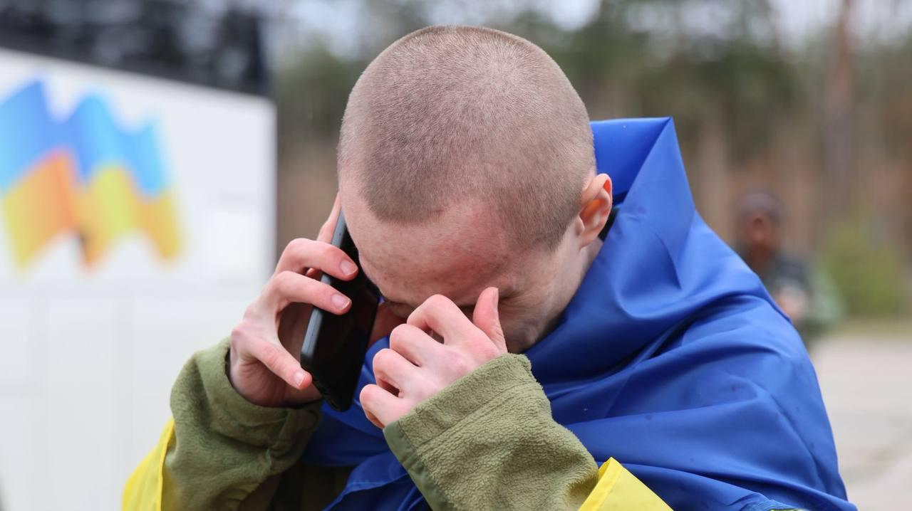 Український захисник, огорнутий у синьо-жовтий прапор, емоційно розмовляє по телефону, прикриваючи обличчя рукою.