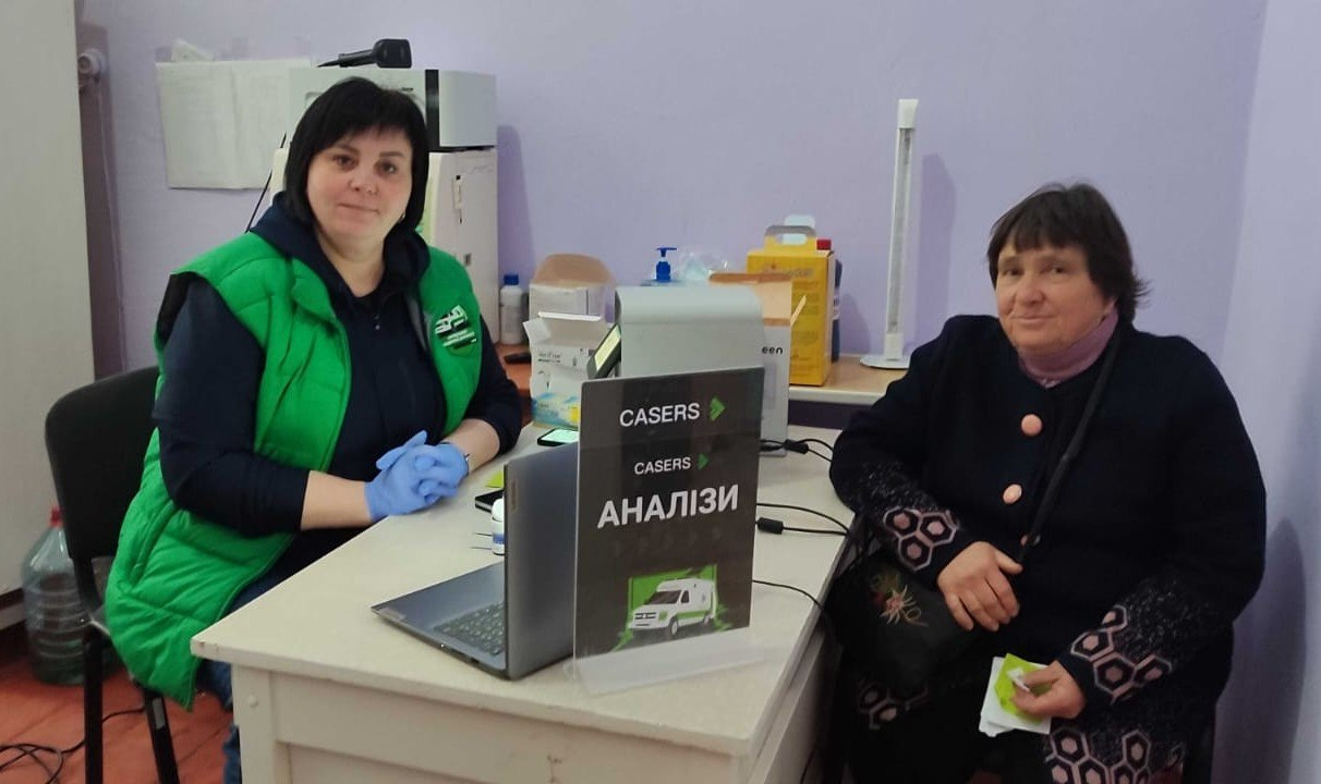 Медичний кабінет: працівниця у зеленому жилеті за столом з ноутбуком приймає пацієнтку. На столі табличка з написом «CASERS АНАЛІЗИ».