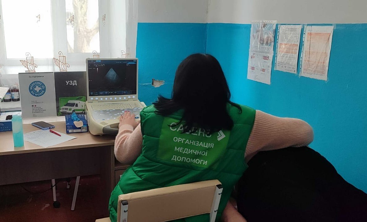 Медична працівниця в зеленому жилеті «CASERS» проводить УЗД-обстеження пацієнту в кабінеті.