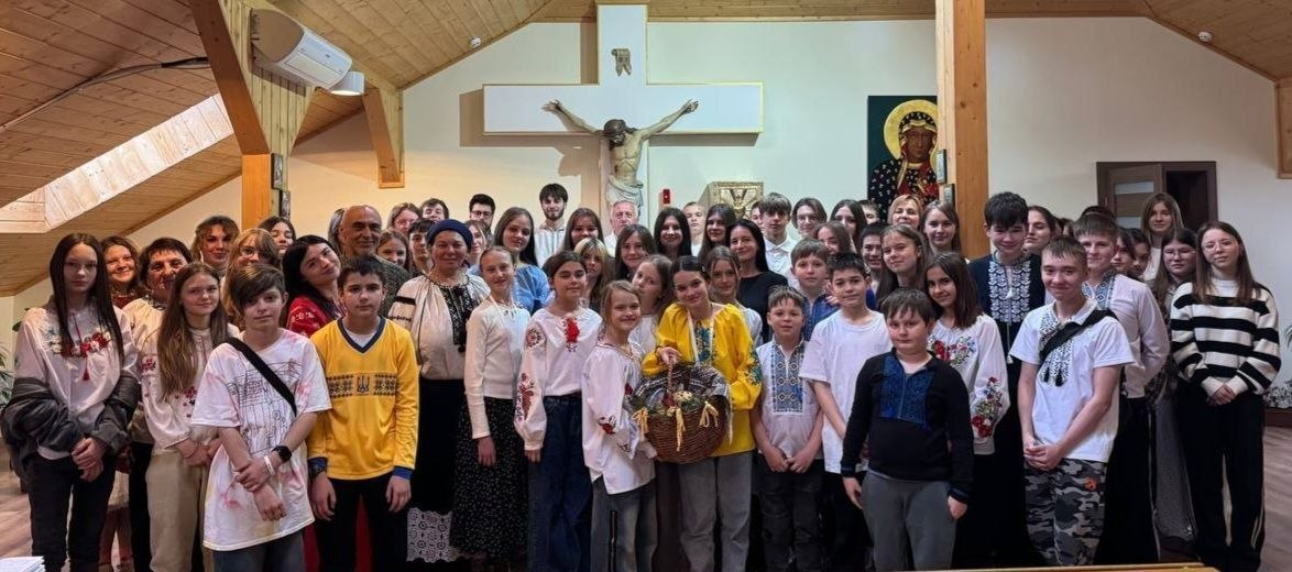 Група молоді та дорослих у приміщенні з дерев'яним оздобленням, багато хто в традиційному українському вбранні — вишиванках. У центрі дівчина тримає плетений кошик. На задньому плані видно велике розп'яття та ікону.