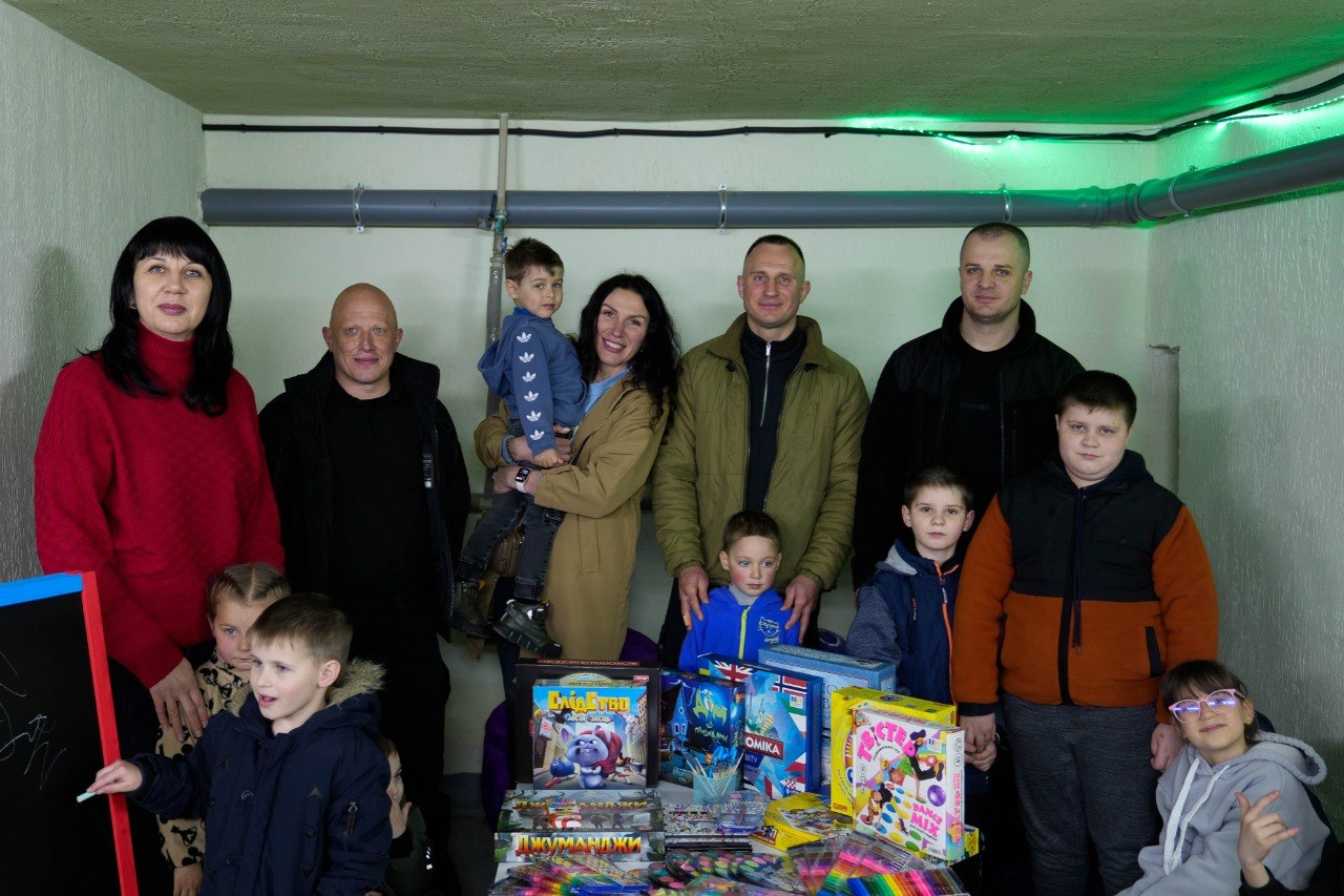 Група дорослих і дітей у приміщенні поруч із наборами іграшок, настільних ігор та приладдям для малювання.