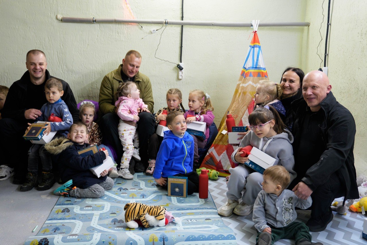 Група дорослих та дітей у дитячій ігровій кімнаті з подарунками в руках. Діти сидять на ігровому килимку поруч із іграшковим наметом-вігвамом.