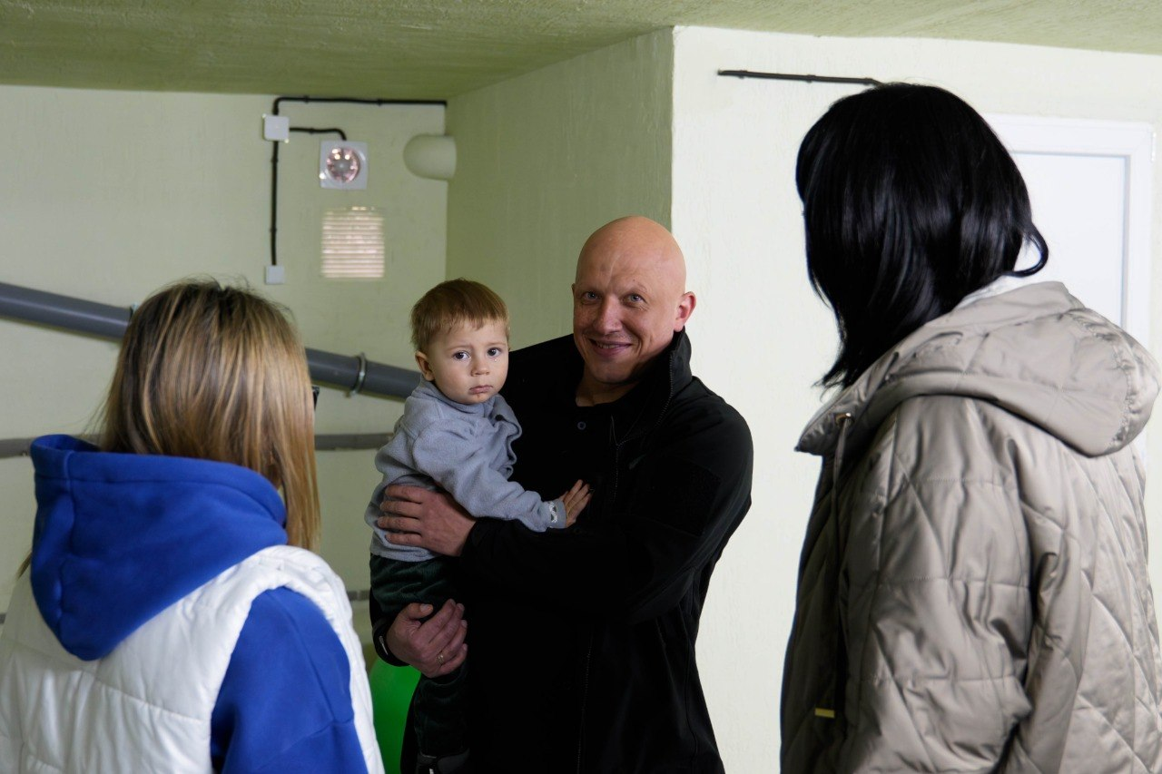 Чоловік тримає на руках маленьку дитину в оточенні двох жінок у приміщенні зі світлими стінами.