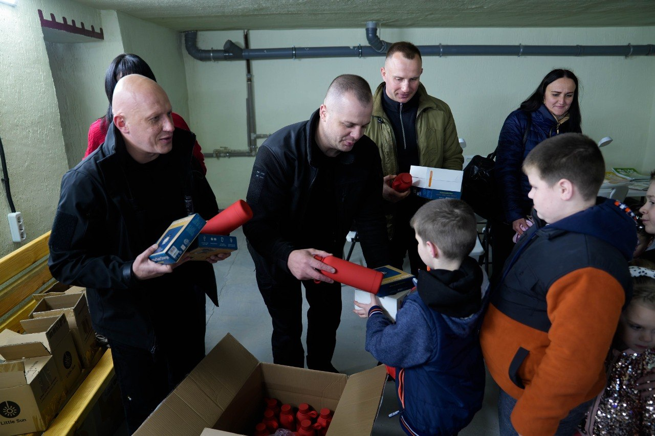 Група дорослих роздає дітям подарунки з картонної коробки в облаштованому приміщенні.