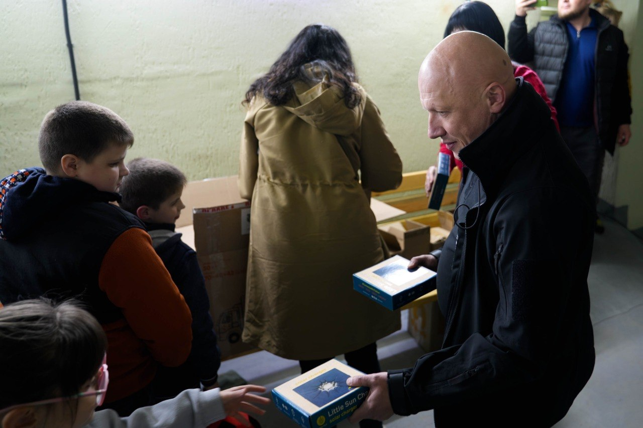 Чоловік передає дітям сині коробки з написом «Little Sun Charge» біля картонних пакунків у приміщенні.