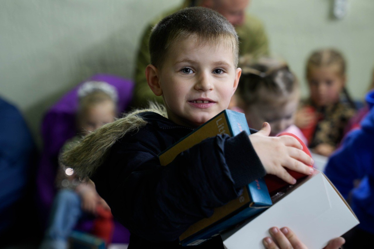 Хлопчик тримає в руках кілька коробок із подарунками на фоні інших дітей у приміщенні.