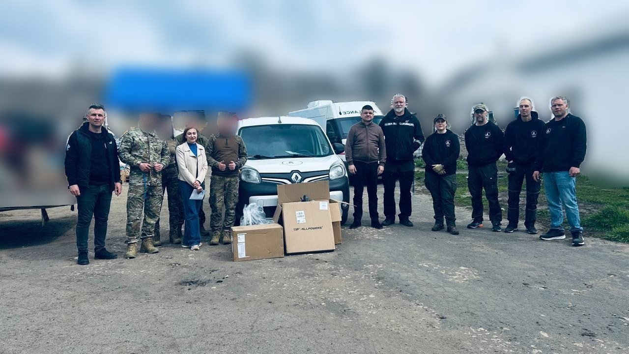 Група людей у цивільному та військовому одязі стоїть поруч із білим фургоном Renault та картонними коробками на відкритій місцевості.