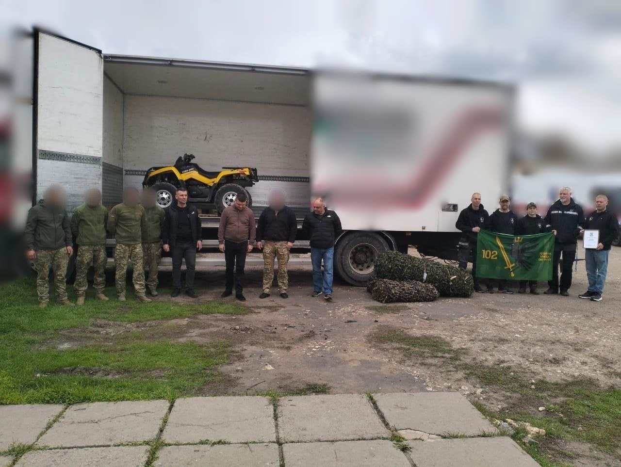 Група людей у цивільному та військовому одязі стоїть поруч із вантажівкою, у кузові якої знаходиться жовтий квадроцикл. Праворуч троє осіб тримають зелений прапор із написом «102».