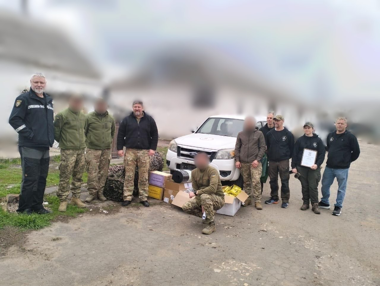 Група людей у цивільному та військовому одязі стоїть поруч із білим позашляховиком Ford, картонними коробками та пакунками. Одна з учасниць тримає в руках подяку або сертифікат у рамці.