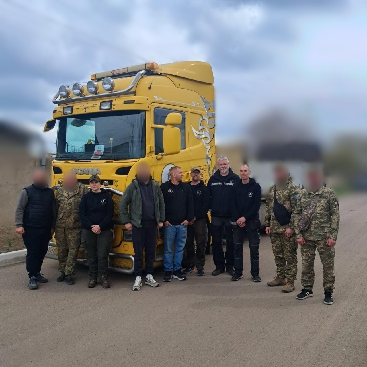 Група людей у цивільному та військовому одязі стоїть на фоні великої жовтої вантажівки Scania на відкритій місцевості.