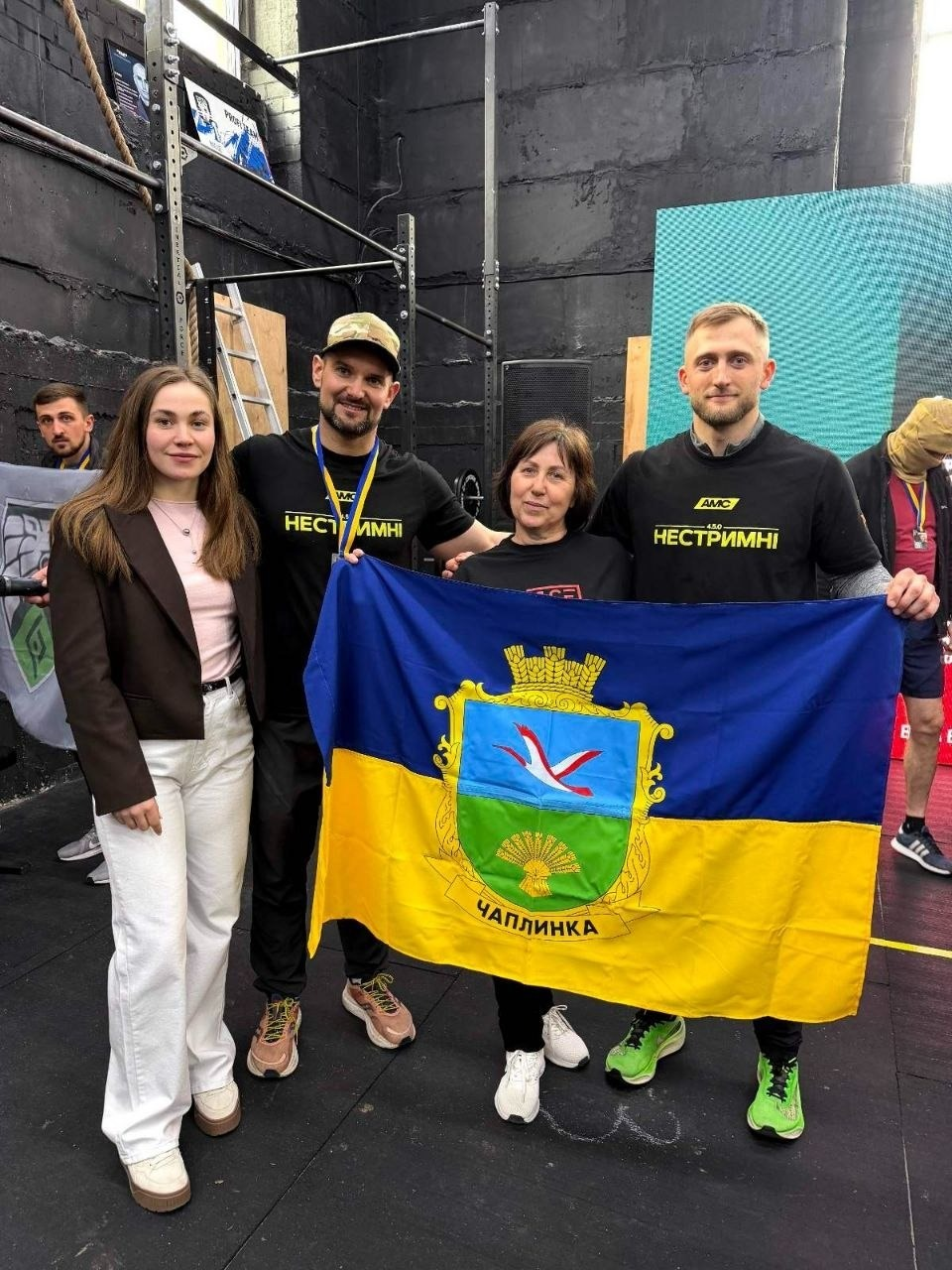 Чотири людини стоять у спортивному залі та тримають синьо-жовтий прапор із гербом і написом «Чаплинка». Чоловіки одягнені в чорні футболки з написом «Нестримні».