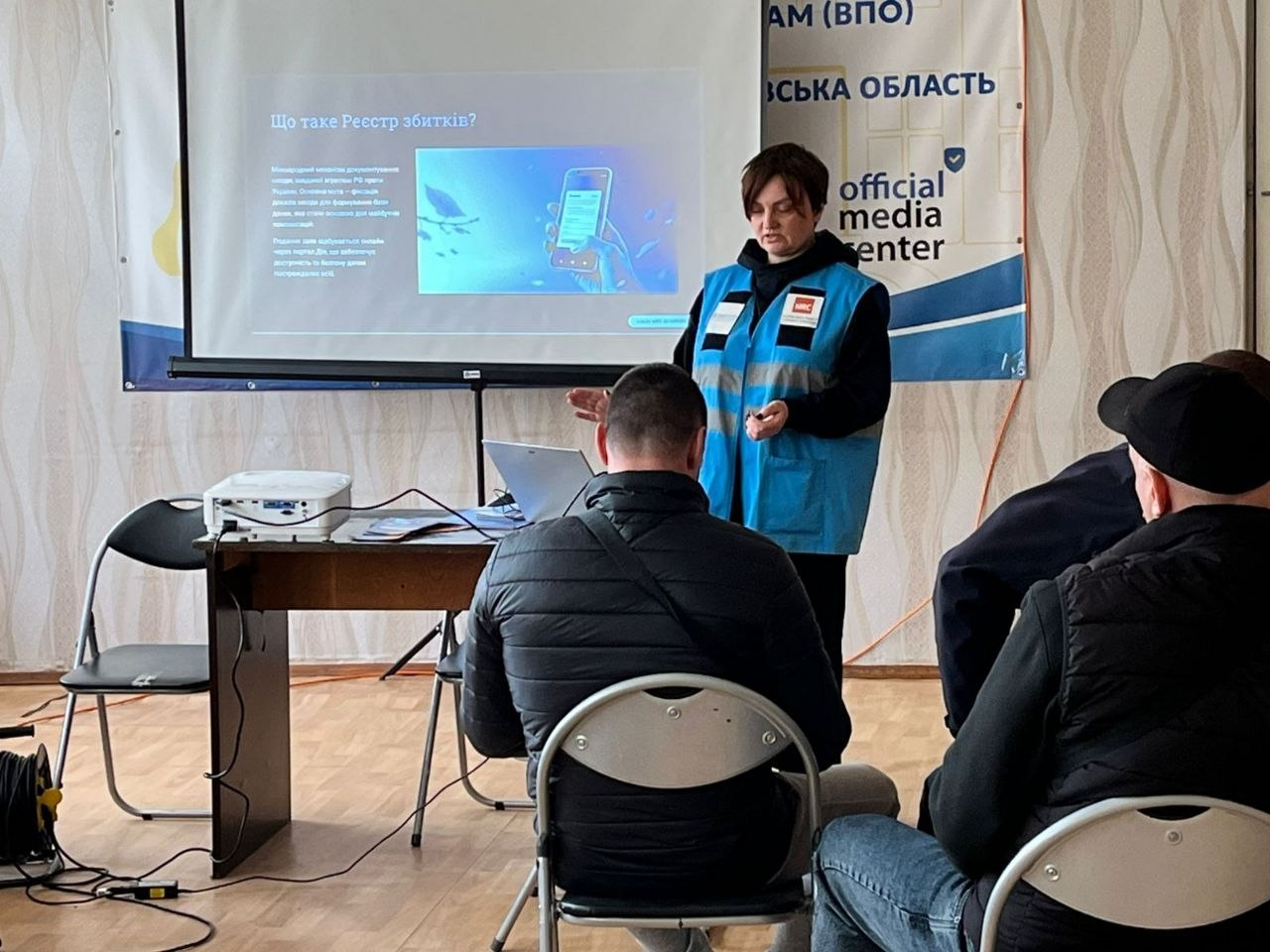 Представниця гуманітарної організації в синьому жилеті проводить презентацію на тему «Що таке Реєстр збитків?» для групи людей у приміщенні медіацентру.