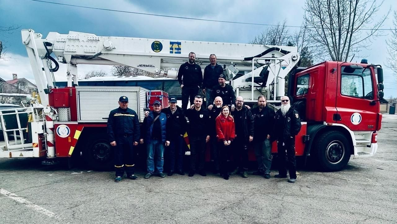 Група людей у цивільному та службовому одязі стоїть поруч із великим червоно-білим пожежним автомобілем із підйомним механізмом на відкритій місцевості.