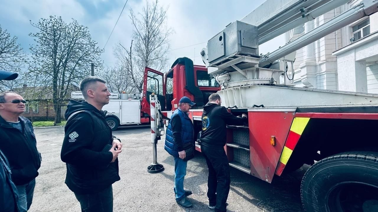Група чоловіків у цивільному одязі оглядає червоно-білий пожежний автомобіль із підйомним механізмом на відкритому майданчику.