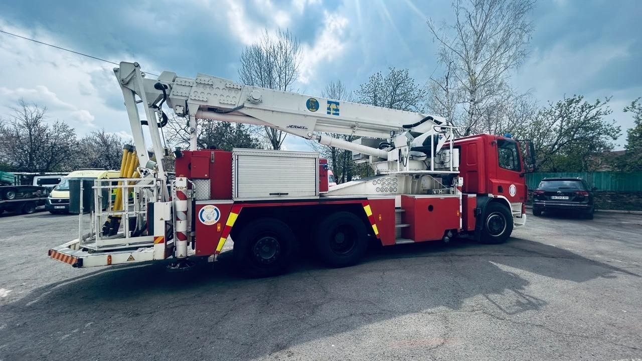Червоно-білий пожежний автомобіль із розкладеним підйомним механізмом на асфальтованому майданчику на фоні дерев та неба.