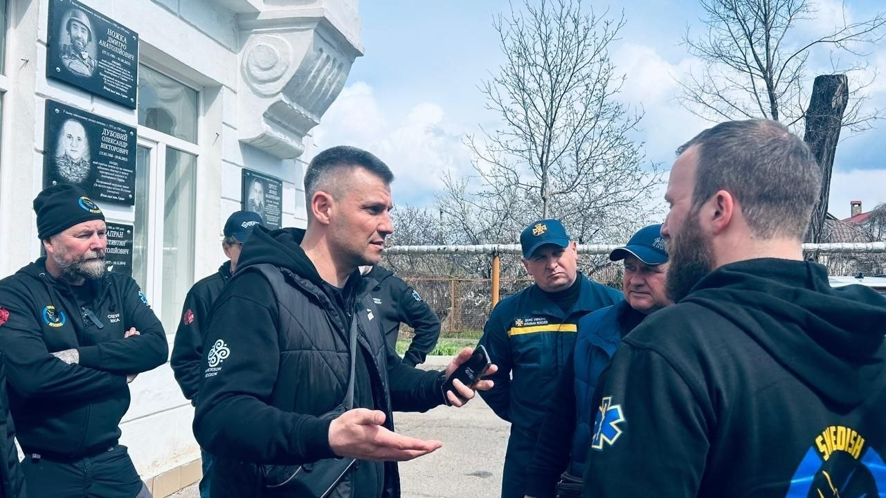 Група чоловіків у темному одязі та формених кепках спілкується на вулиці біля будівлі з пам'ятними дошками на стіні. Один із чоловіків жестикулює, тримаючи в руці смартфон.