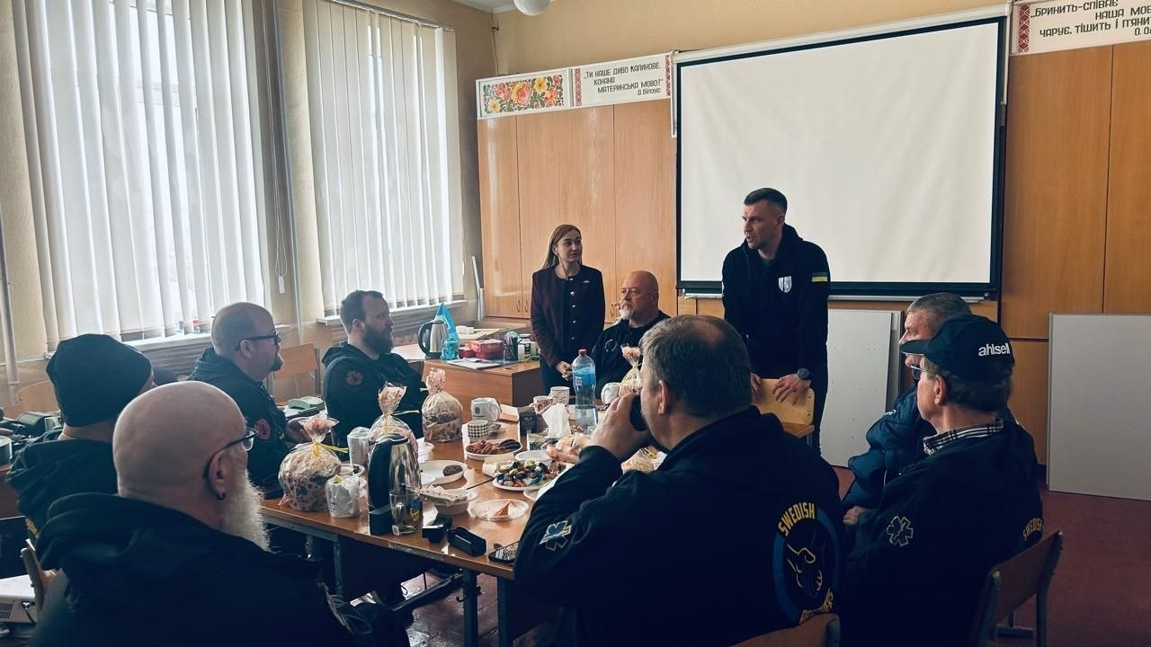 Група людей сидить за довгим столом у приміщенні з великим білим екраном. На столі стоять чашки, тарілки з їжею та паски в упаковках. Один чоловік стоїть і виступає перед присутніми.