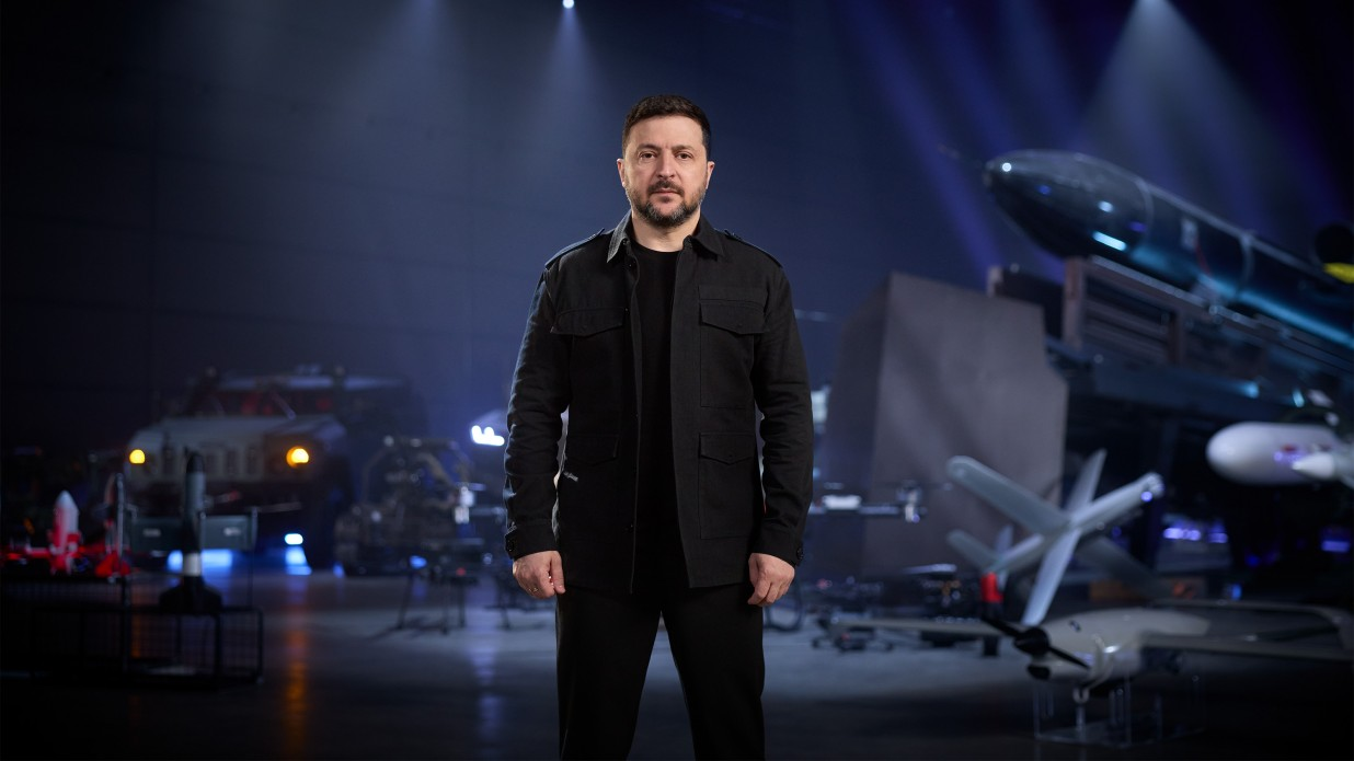 Чоловік у чорному одязі стоїть у центрі ангара на фоні зразків військової техніки, зокрема безпілотників та ракет.