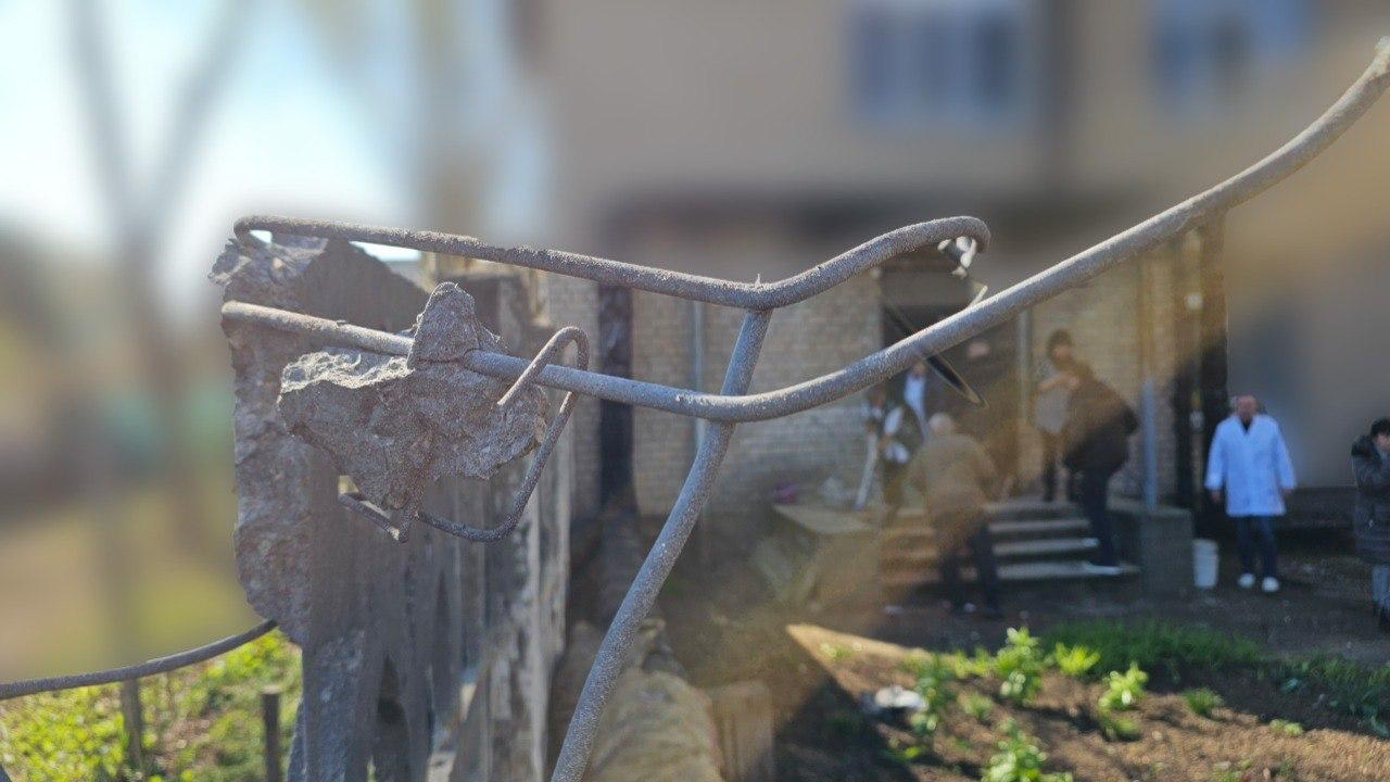 Зруйнований залізобетонний паркан із покрученою арматурою на фоні будівлі, біля якої знаходяться люди.