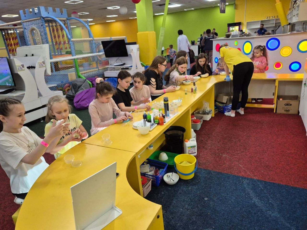 Діти та дорослі за довгим жовтим столом у дитячому центрі беруть участь у майстер-класі з виготовлення слаймів.