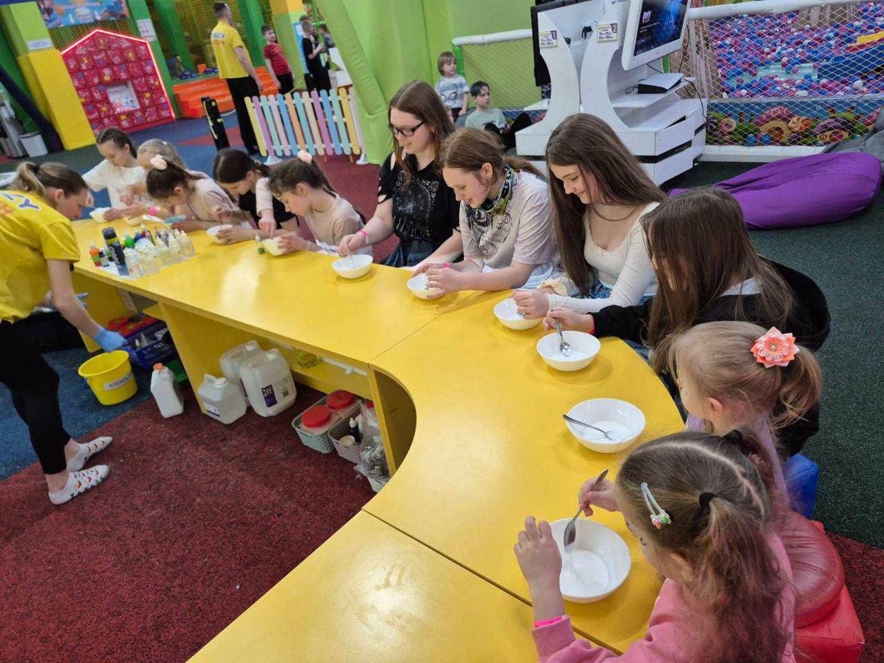 Діти та підлітки за круглим жовтим столом у дитячому центрі займаються творчістю, перемішуючи інгредієнти в білих мисках.