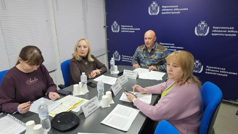 Четверо людей сидять за робочим столом під час наради в офісному приміщенні з синім брендованим фоном.