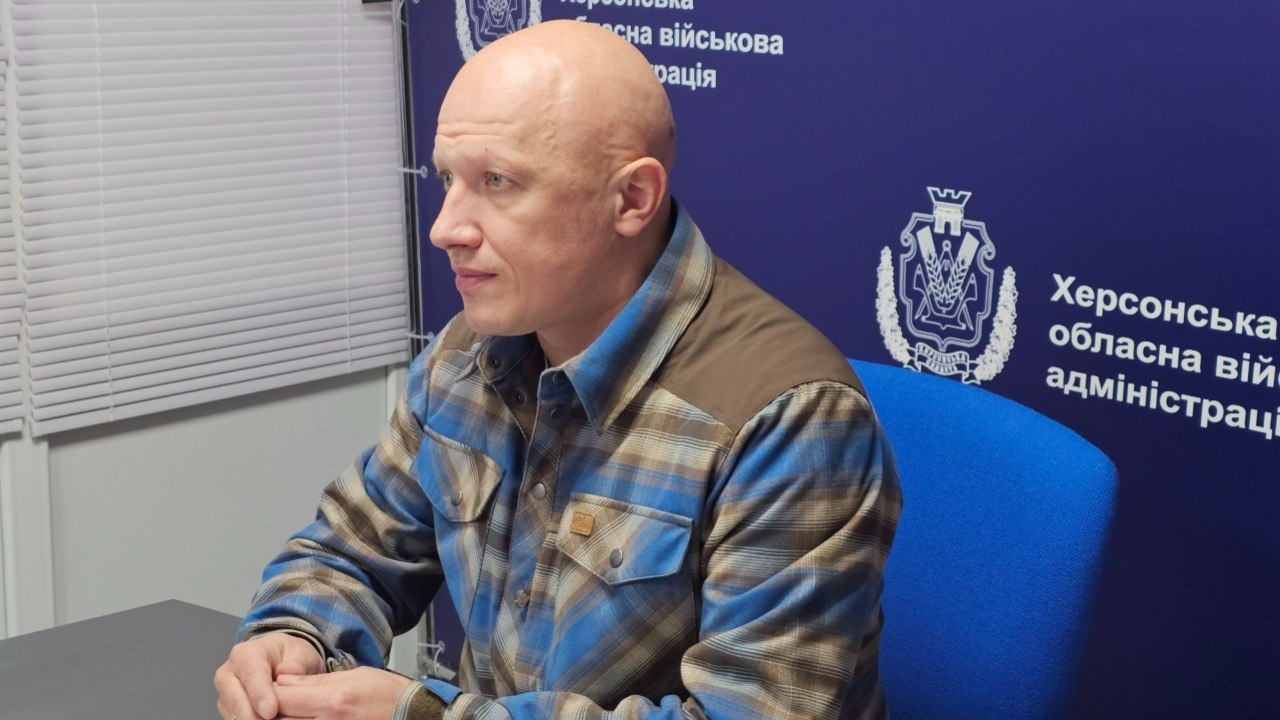 Чоловік у картатій сорочці сидить за столом на фоні синього банера з офіційною символікою.