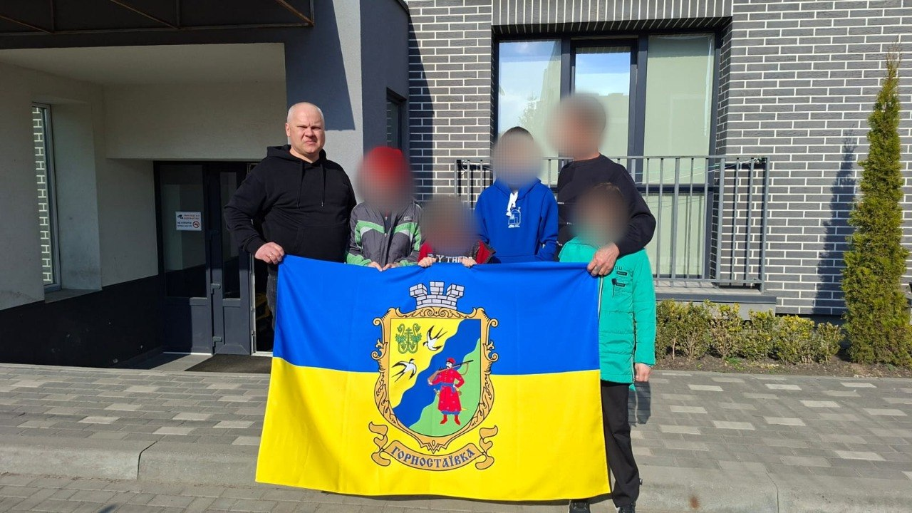 Група людей тримає прапор України з гербом Горностаївки на фоні сучасної будівлі.