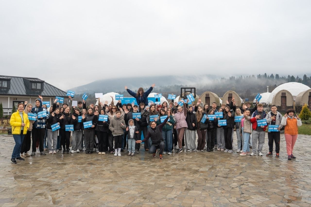 Велика група молоді на площі просто неба тримає в руках однакові сині таблички на фоні гір та купольних споруд.