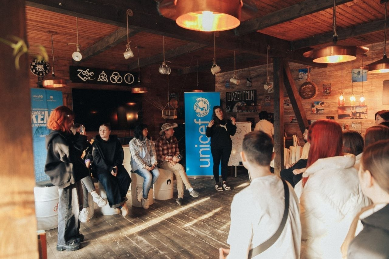 Група молоді на тренінгу в автентичному приміщенні з дерев'яним оздобленням та банером UNICEF на фоні.