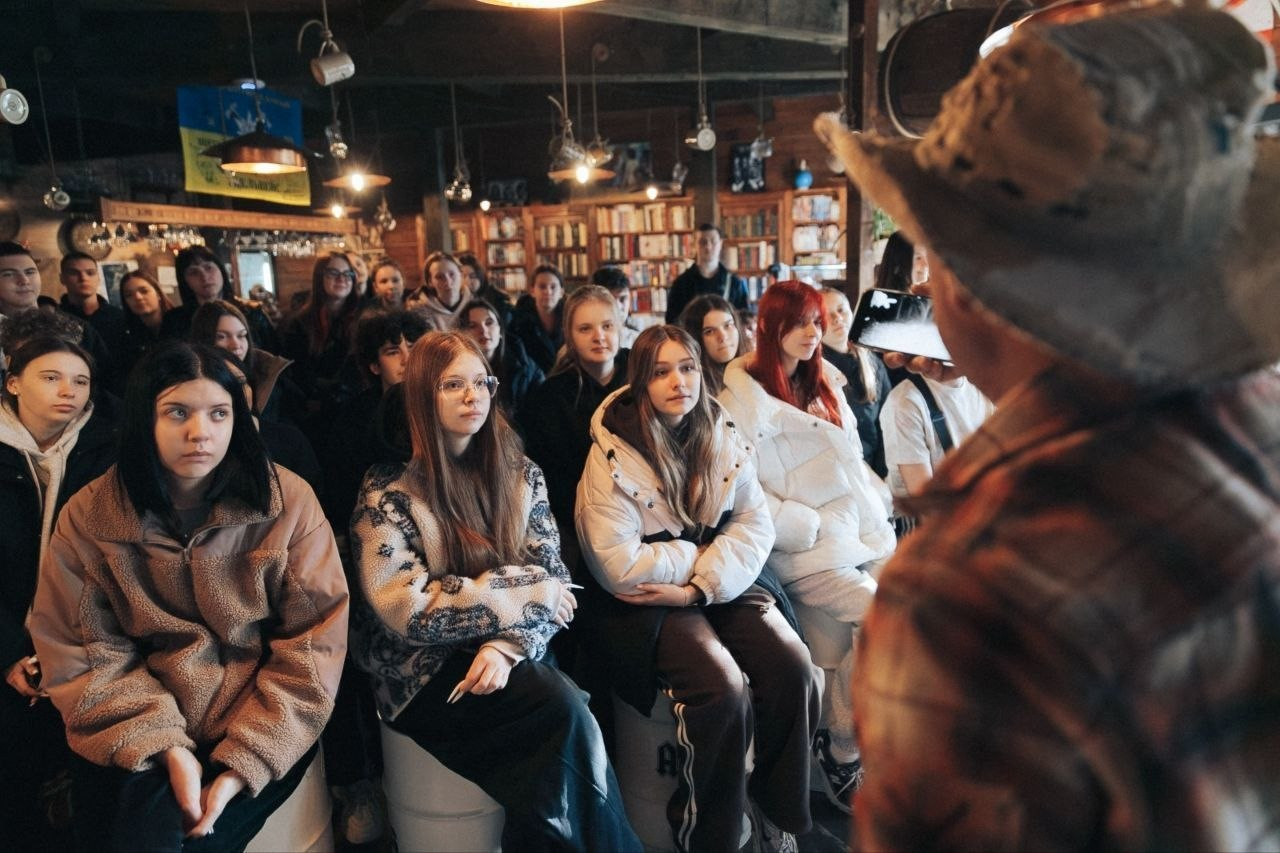 Група молоді в затишній залі з книжковими полицями уважно слухає виступ спікера.