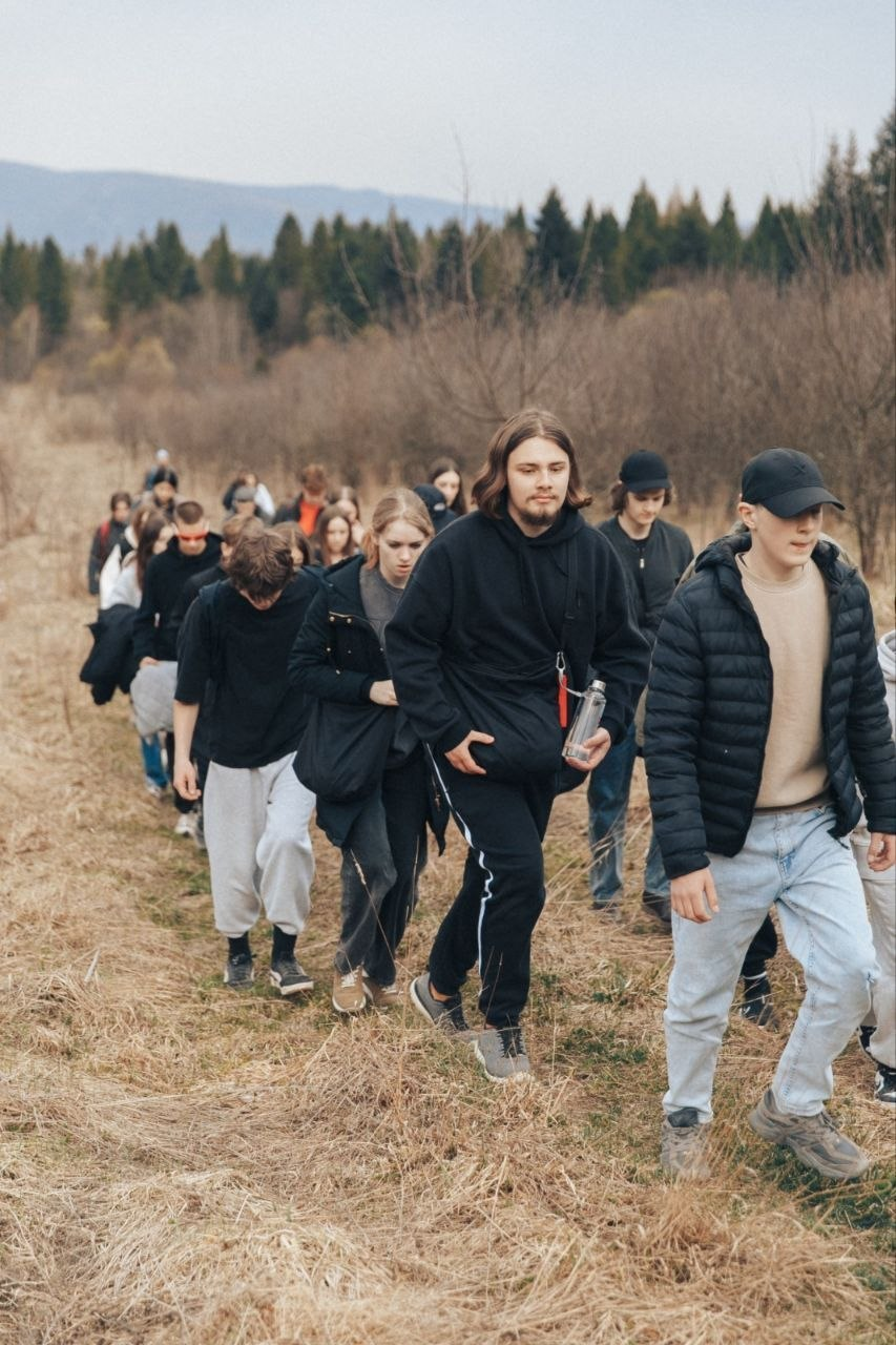 Група молоді йде колоною по трав'янистому схилу на фоні лісу та гір.