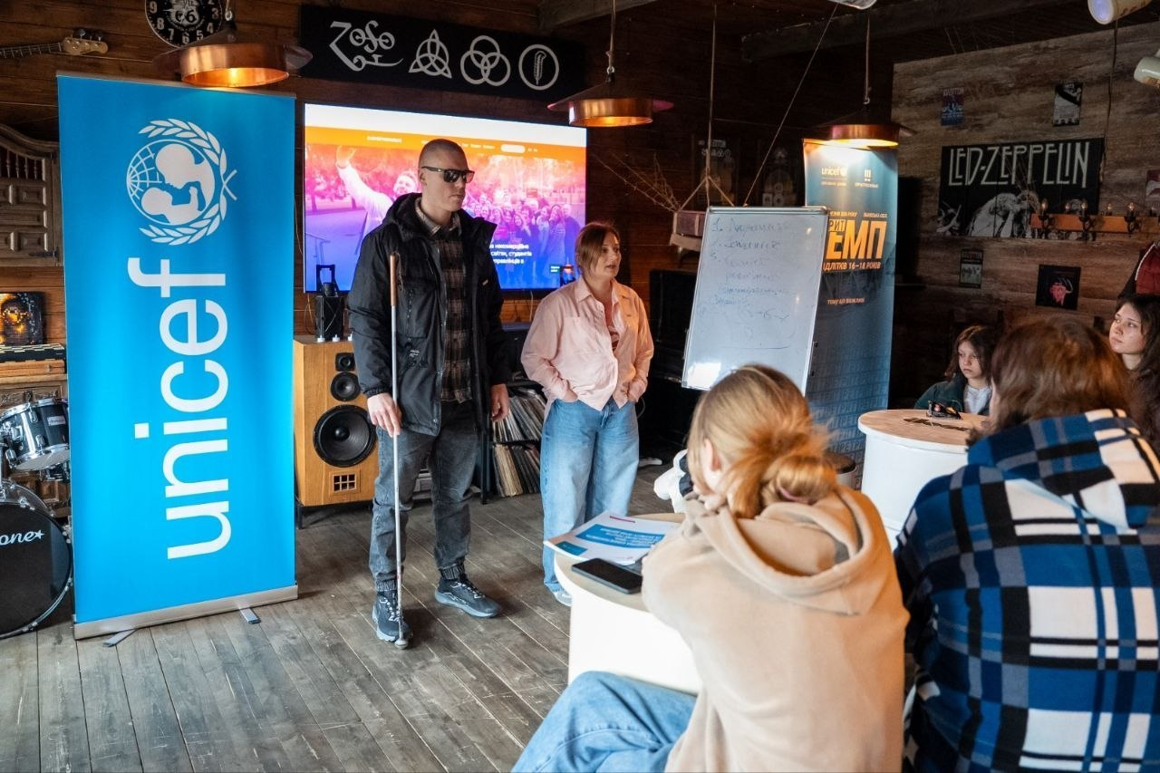 Виступ спікерів перед аудиторією в автентичному залі з дерев'яним оздобленням, телевізором та великим банером UNICEF.