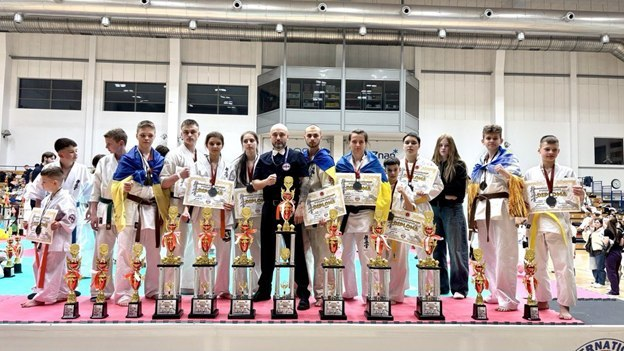 Група спортсменів у кімоно з кубками, дипломами та прапорами України стоїть на п'єдесталі під час нагородження на змаганнях з карате.