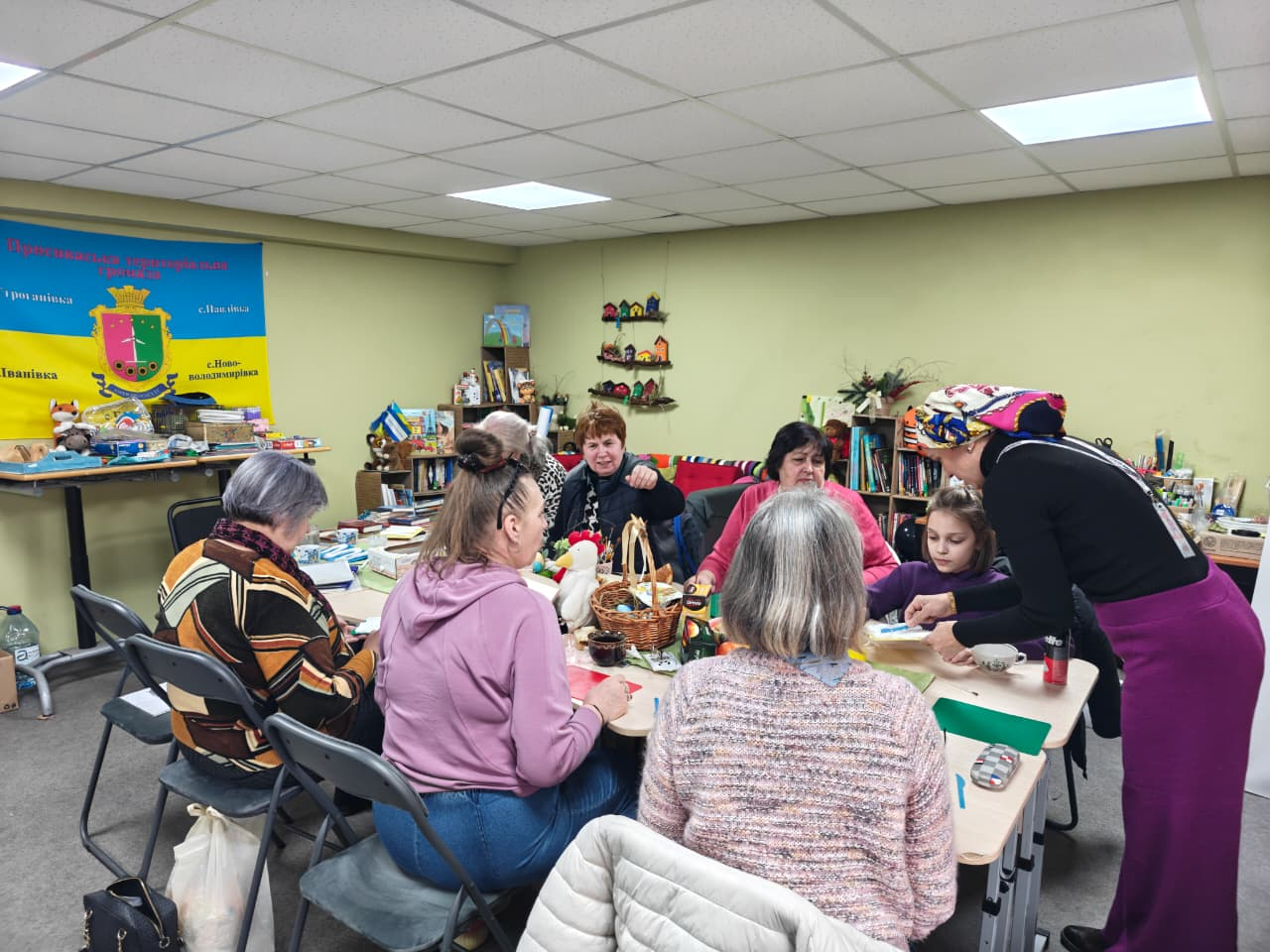 Група жінок та дитина займаються рукоділлям за великим столом у затишному приміщенні з книжковими полицями та патріотичною символікою на стіні.