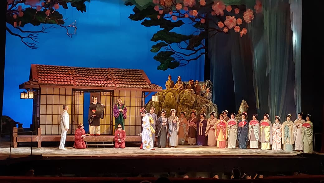 Сцена з театральної вистави в японському стилі з акторами в традиційних костюмах на фоні декорацій будинку та квітучих дерев.