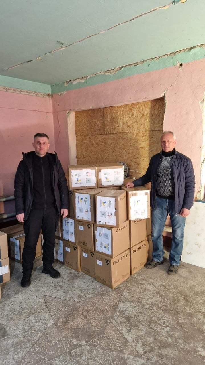 Двоє чоловіків стоять біля купи картонних коробок з гуманітарною допомогою та обладнанням Bluetti в обшарпаному приміщенні.