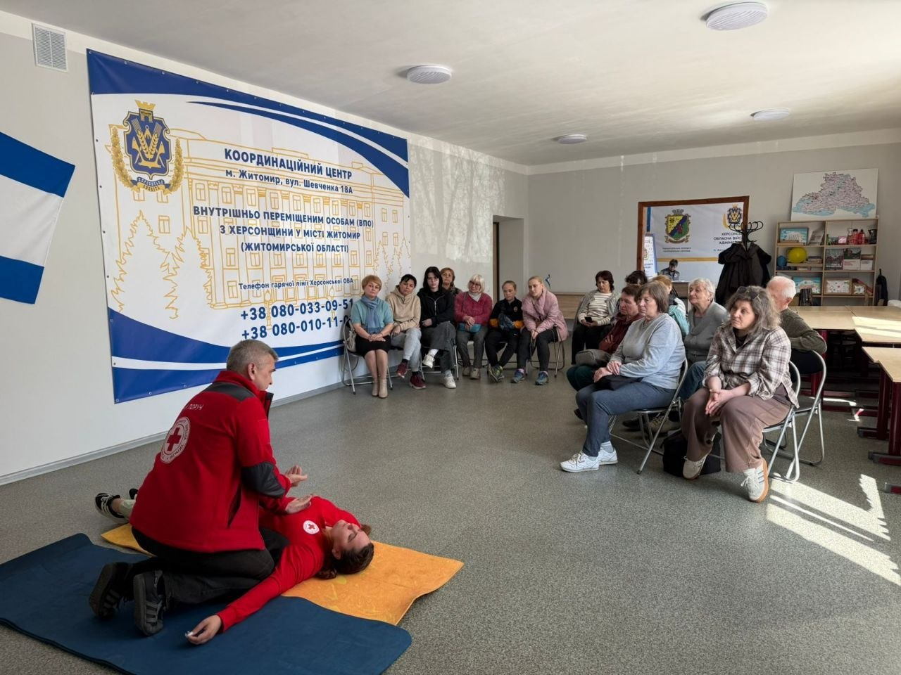 Інструктор Червоного Хреста проводить тренінг із домедичної допомоги для групи людей у координаційному центрі. На передньому плані демонструють техніку допомоги людині, що лежить на килимку.