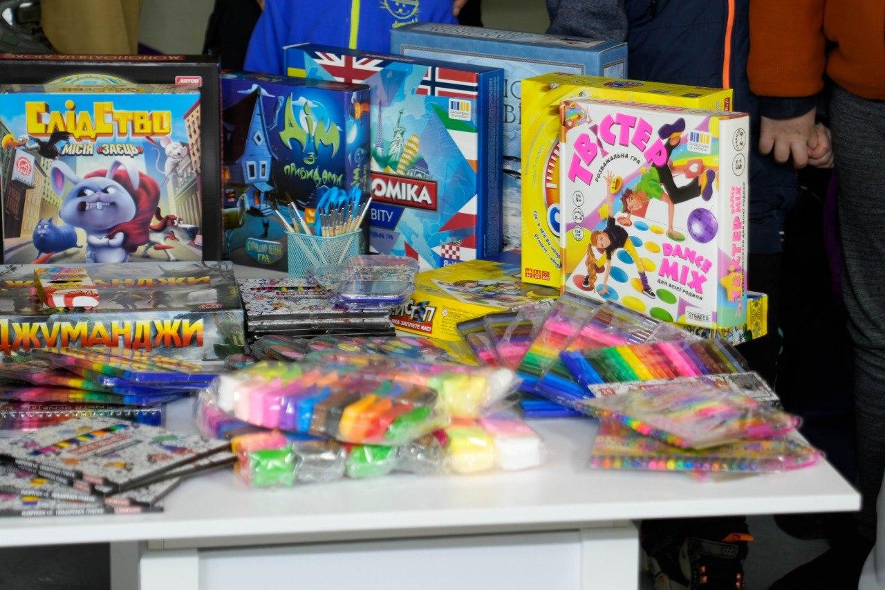 На столі розкладено різноманітні дитячі товари: настільні ігри в яскравих коробках, набори пластиліну, фломастери та інше приладдя для творчості.