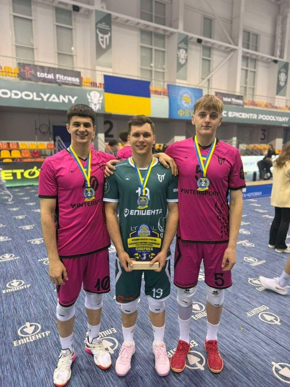 Три волейболісти в ігровій формі з медалями та нагородою стоять на спортивній арені після завершення змагань.