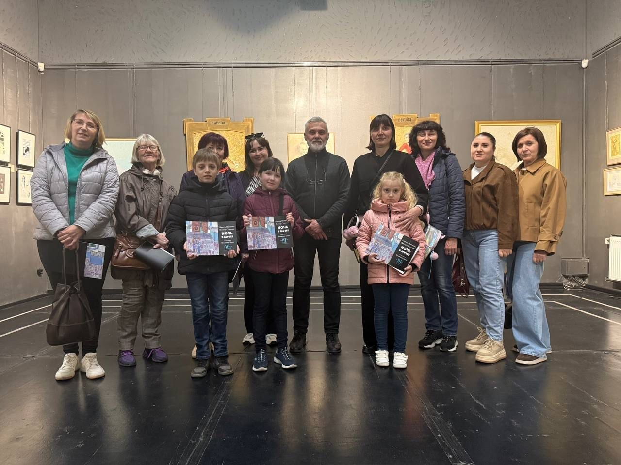 Група дорослих і дітей стоїть у музейній залі на фоні картин. Діти тримають у руках пам'ятні грамоти або буклети з написом «Ніч у музеї».