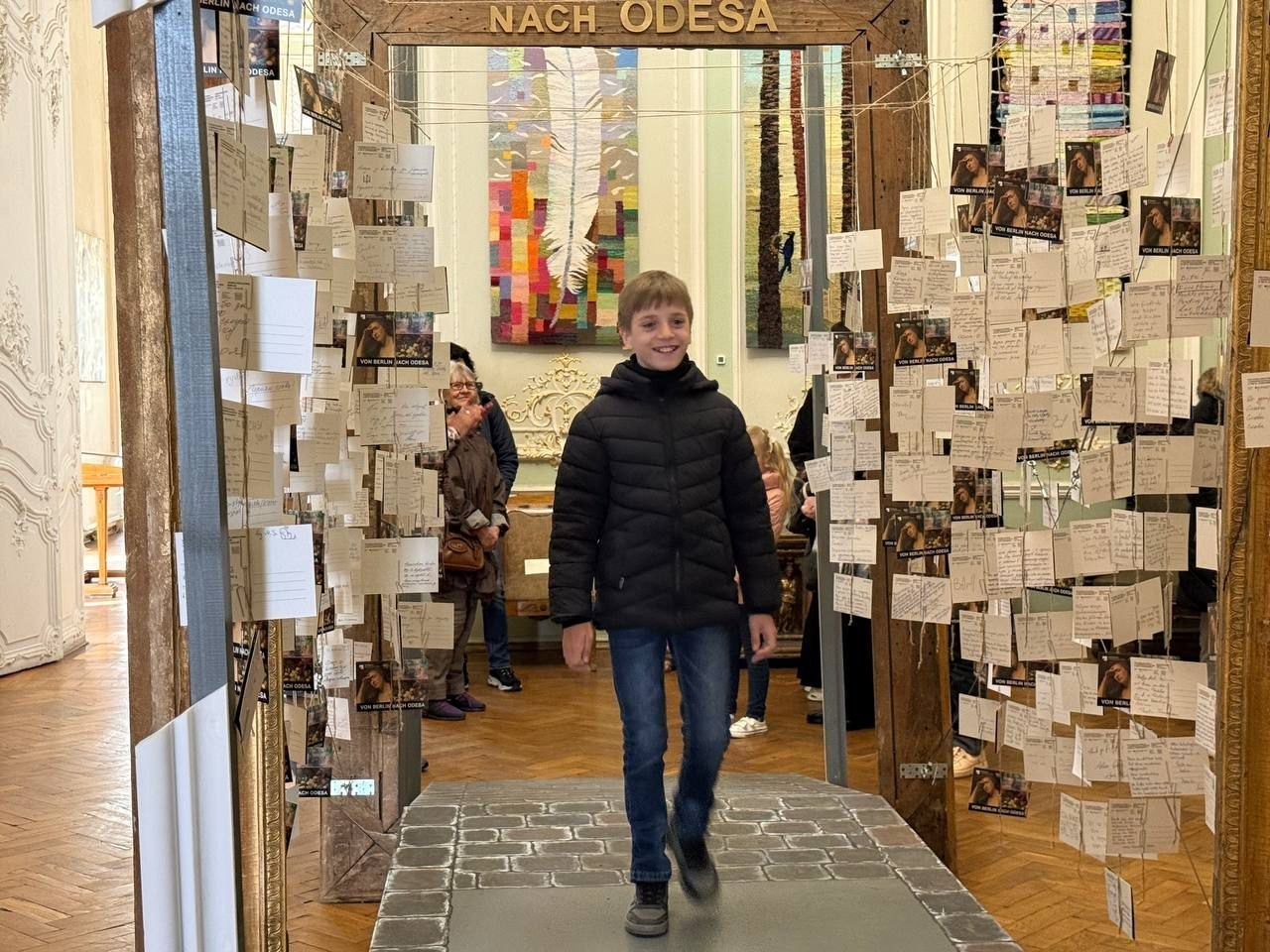 Хлопчик проходить крізь дерев’яну арку, прикрашену численними картками з текстом та фотографіями, у музейному або виставковому залі. На фоні видно гобелен та інших відвідувачів.