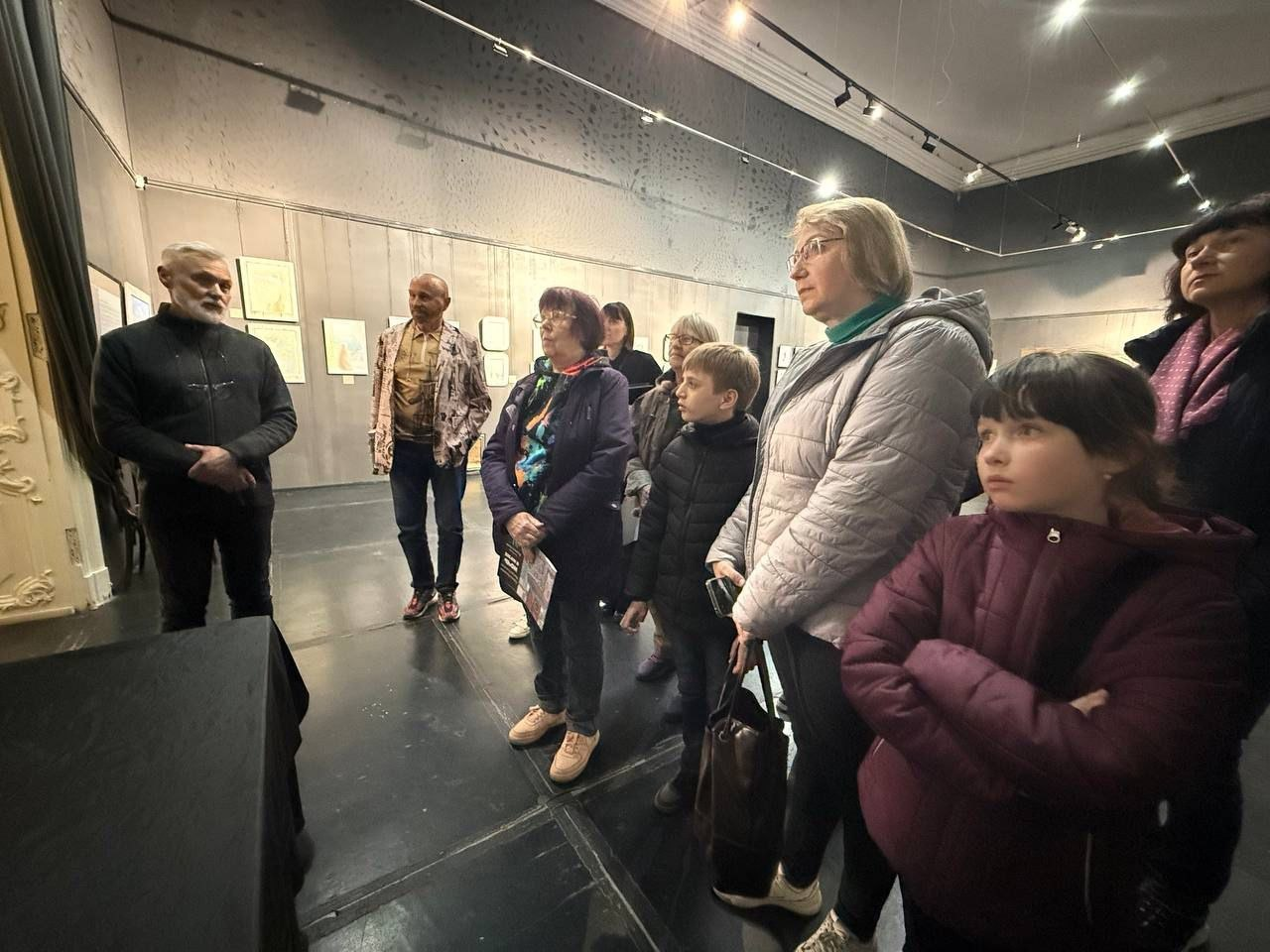 Група відвідувачів, серед яких дорослі та діти, слухають екскурсовода в музейній залі з картинами на стінах.