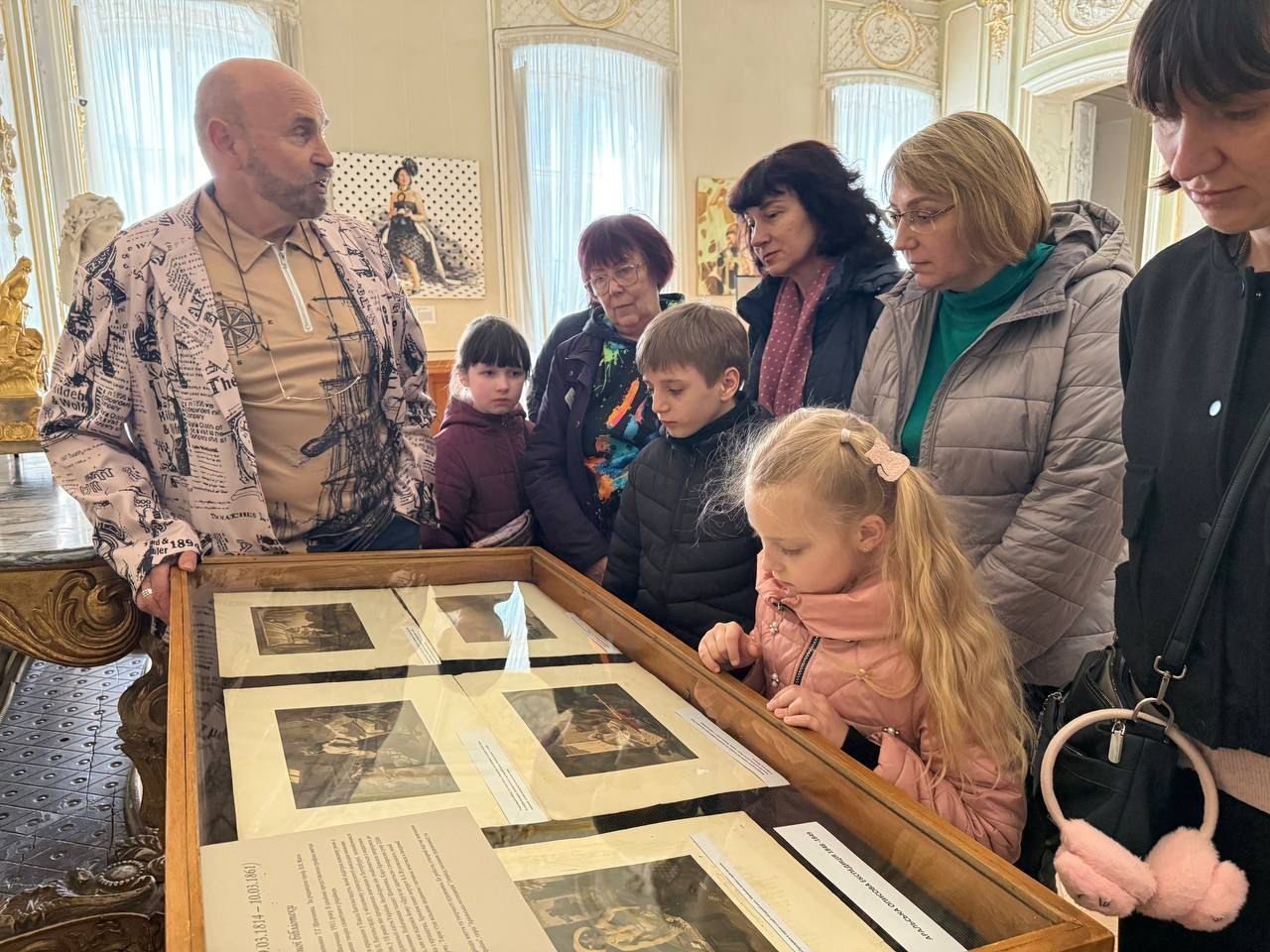 Група людей, серед яких діти та дорослі, розглядають експонати в заскленій вітрині музею під час екскурсії.