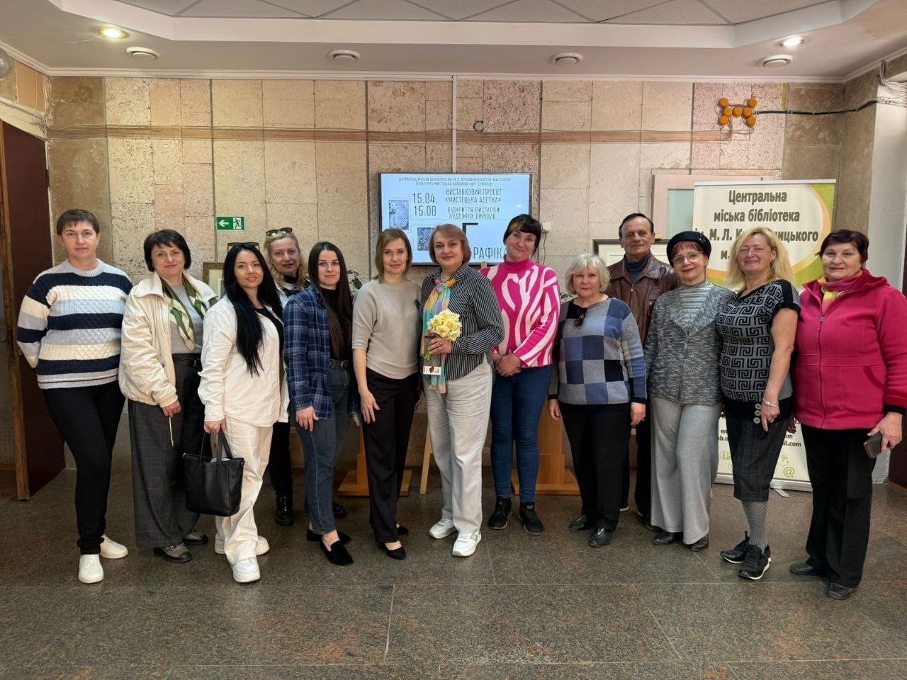 Групове фото людей у холі бібліотеки. Учасники стоять перед інформаційними стендами та афішами заходів.