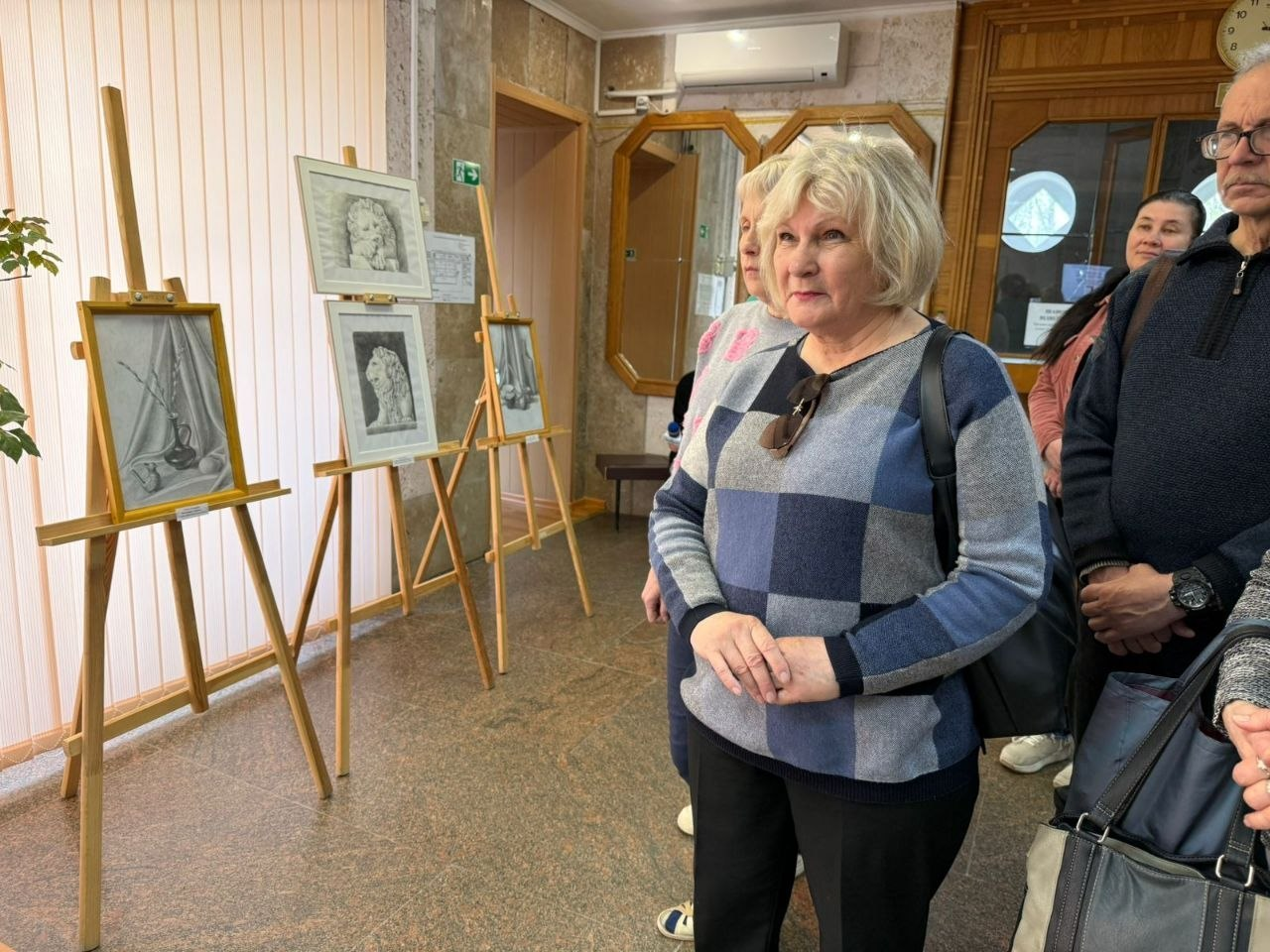 Відвідувачі розглядають виставку графічних робіт, розміщених на дерев'яних мольбертах у світлому приміщенні.