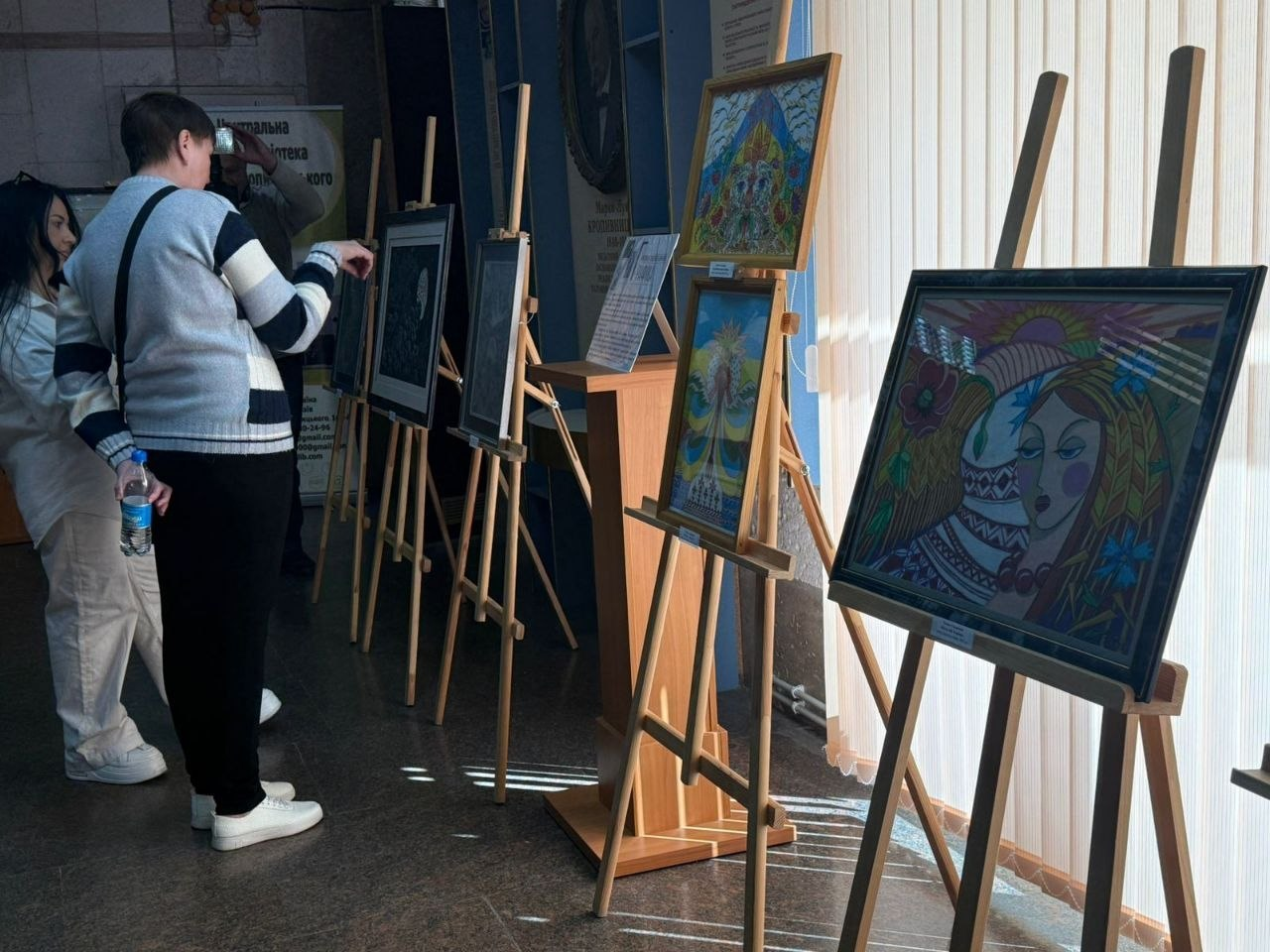 Відвідувачі оглядають виставку картин у приміщенні бібліотеки. Твори у рамах розміщені на дерев'яних мольбертах вздовж вікна.