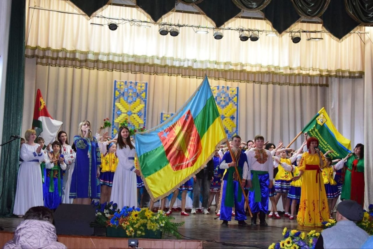 Група людей у традиційному українському вбранні виступає на сцені. У центрі композиції чоловік тримає великий синьо-жовто-зелений прапор із гербом. На фоні сцени розміщені декорації з національними орнаментами та світле драпірування.