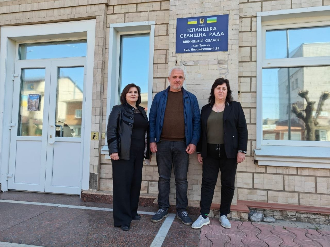 Троє людей — дві жінки та чоловік — стоять перед входом до будівлі Теплицької селищної ради. Над ними закріплена синя інформаційна табличка з назвою установи та адресою. Будівля має світлий фасад, облицьований плиткою, та великі вікна.